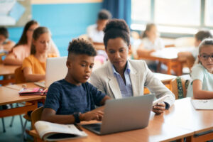 Teacher helping student on laptop in full classroom